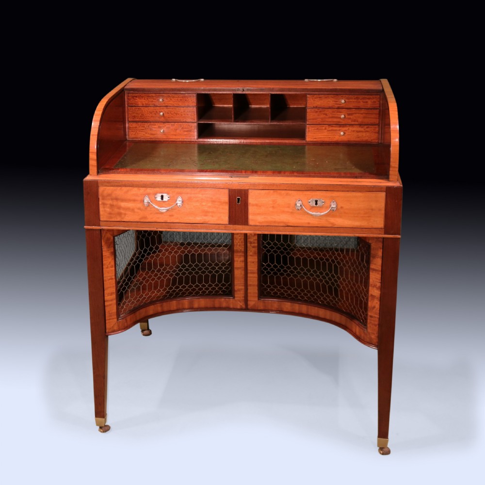 A George III mahogany and satinwood writing table featuring a roll-top compartment, green leather writing surface, inner drawers, and wire mesh lower panels on tapered legs with brass castors. View II