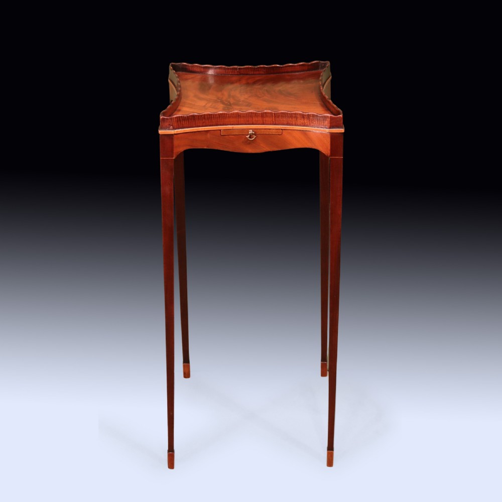 A late 18th-century Sheraton mahogany kettle stand featuring a scalloped gallery top, pull-out slide, and slender square tapered legs.