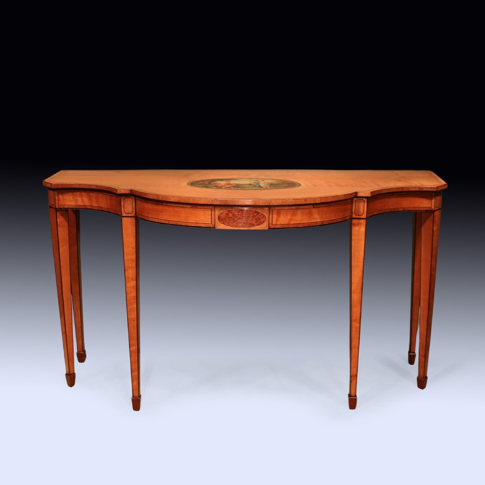 An antique Sheraton period satinwood console table featuring a curved front, hand-painted central medallion on top, crossbanded borders, and four square tapered legs.