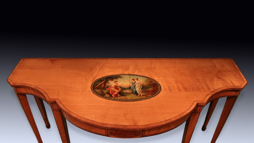 An antique Sheraton period satinwood console table featuring a curved front, hand-painted central medallion on top, crossbanded borders, and four square tapered legs. View VI