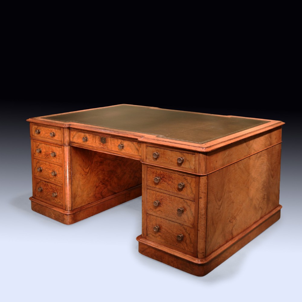 A mid 19th-century Victorian burr walnut pedestal desk featuring a green leather inset top, rounded corners, multiple drawers with turned wooden knobs, and a plinth base.