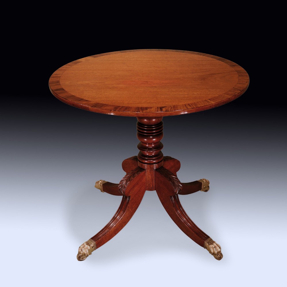 A circular mahogany occasional table featuring a crossbanded top, turned central pedestal, and four splayed legs with brass lion paw feet.