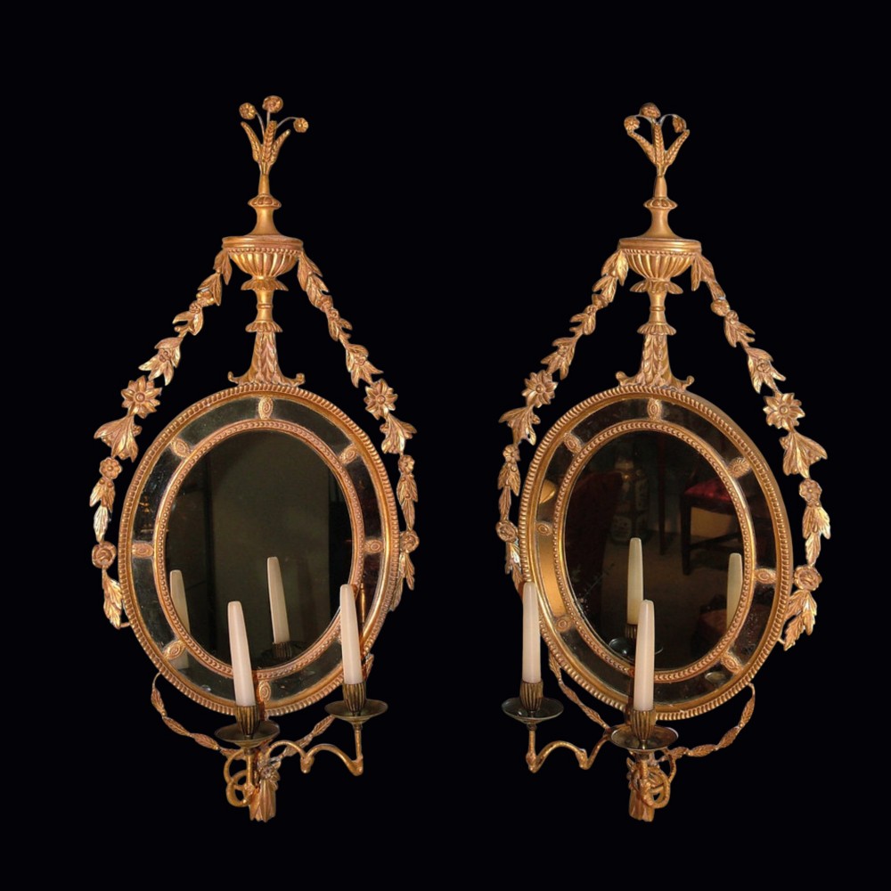 A pair of late 18th-century Neoclassical oval giltwood girandole mirrors with floral swags, urn finials, and twin candle branches.