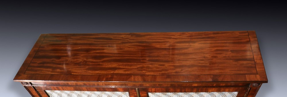 A Regency period two-door mahogany side cabinet featuring brass wire grille panels lined with pleated fabric and raised on turned legs. View IV