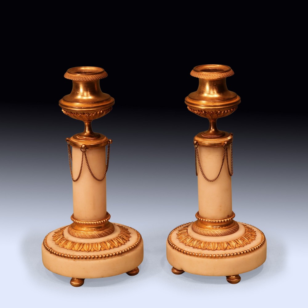 A pair of 19th-century Neoclassical candlesticks featuring white marble cylindrical columns adorned with gilt bronze chain swags and urn-shaped sconces resting on circular bases.