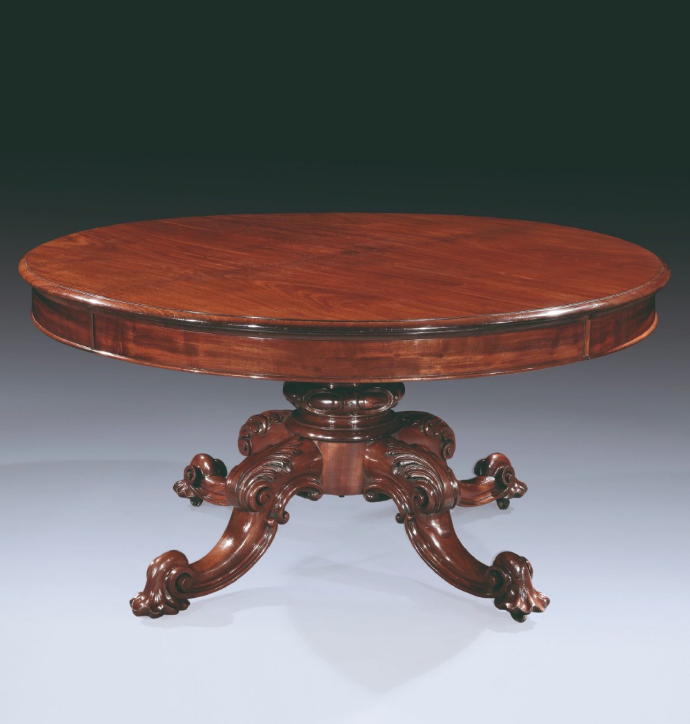 A circular mahogany extending dining table with a thick moulded top resting on a heavily carved central pedestal with four ornate scrolled legs.