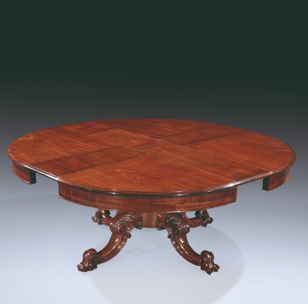 A circular mahogany extending dining table with a thick moulded top resting on a heavily carved central pedestal with four ornate scrolled legs. View II