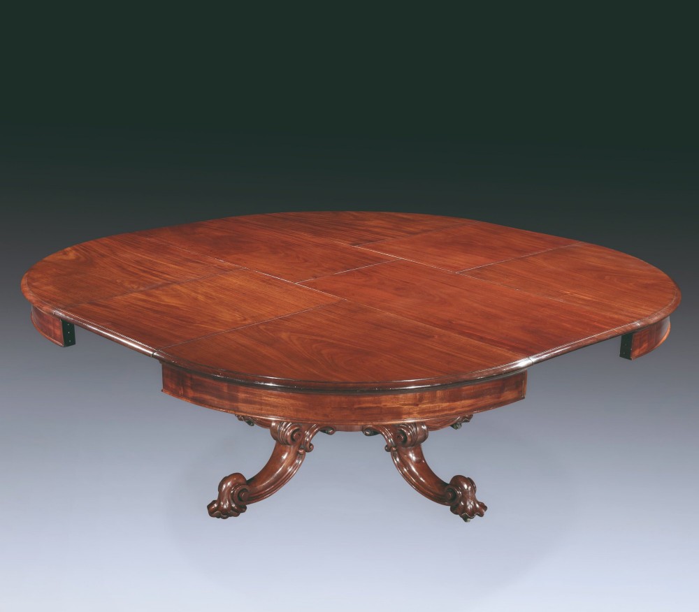 A circular mahogany extending dining table with a thick moulded top resting on a heavily carved central pedestal with four ornate scrolled legs. View III