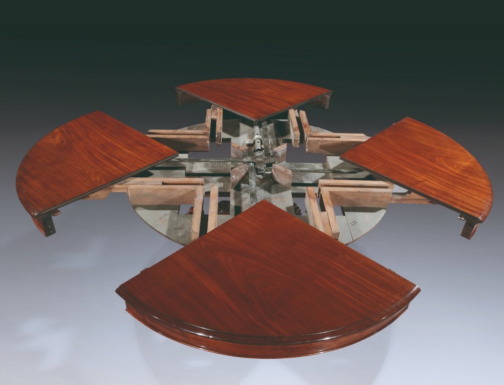 A circular mahogany extending dining table with a thick moulded top resting on a heavily carved central pedestal with four ornate scrolled legs. View IV