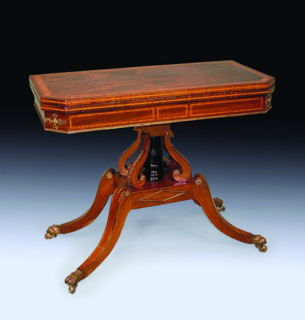 An antique Regency period card table made of dark rosewood with satinwood crossbanding, resting on an intricate scroll pedestal base with four splayed legs ending in brass lion paw castors.