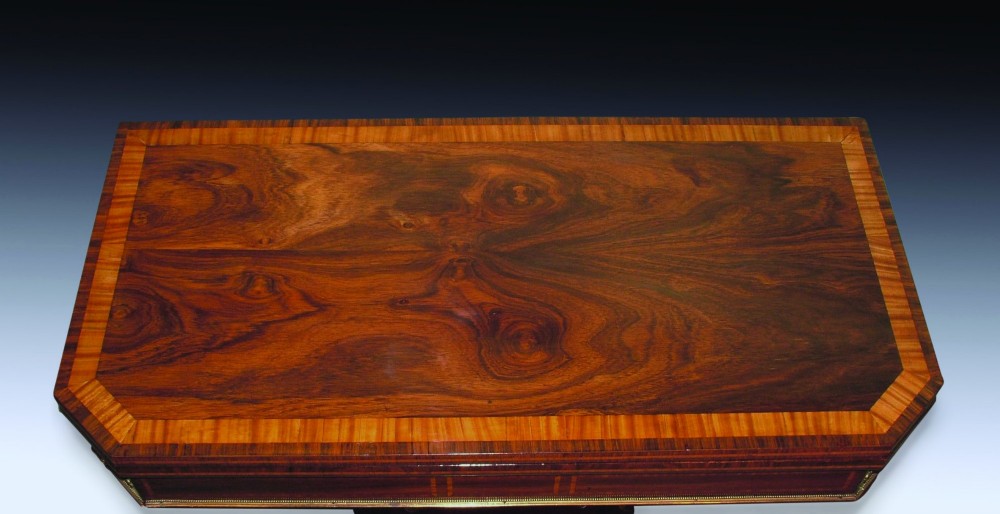 An antique Regency period card table made of dark rosewood with satinwood crossbanding, resting on an intricate scroll pedestal base with four splayed legs ending in brass lion paw castors. View II