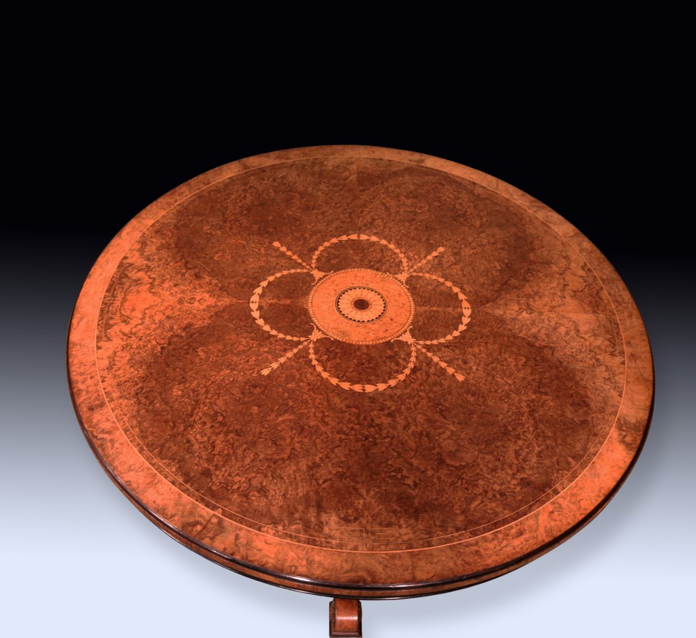 A circular mid 19th-century burr walnut centre table featuring central marquetry inlay and a carved pedestal base with splayed legs. View II