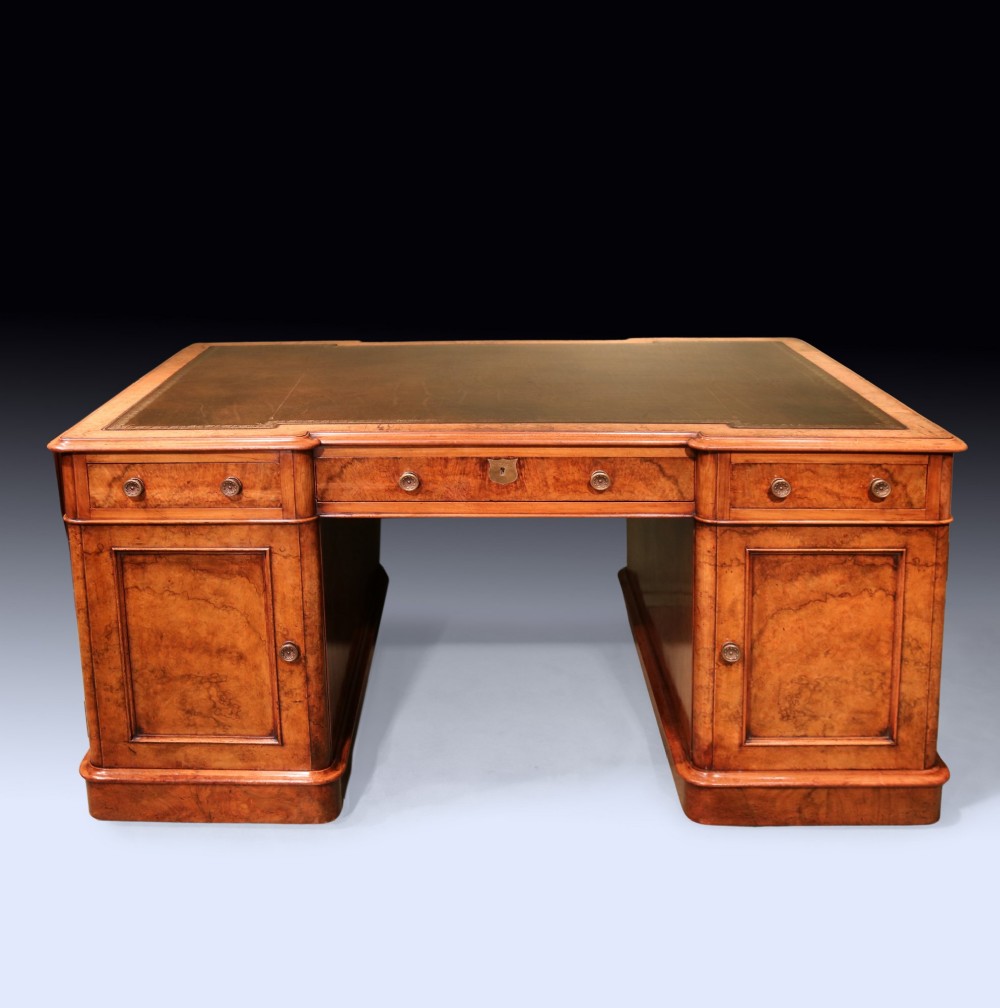 A mid 19th-century Victorian burr walnut pedestal desk featuring a green leather inset top, rounded corners, multiple drawers with turned wooden knobs, and a plinth base. View V