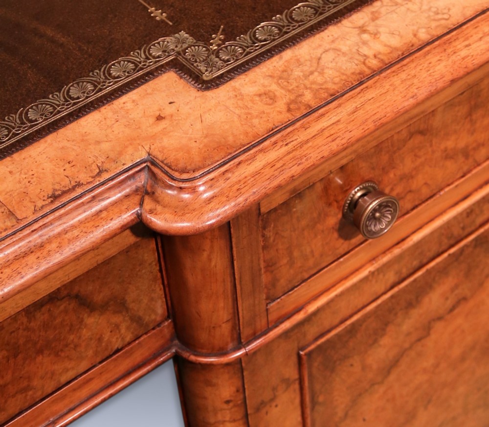 A mid 19th-century Victorian burr walnut pedestal desk featuring a green leather inset top, rounded corners, multiple drawers with turned wooden knobs, and a plinth base. View VI