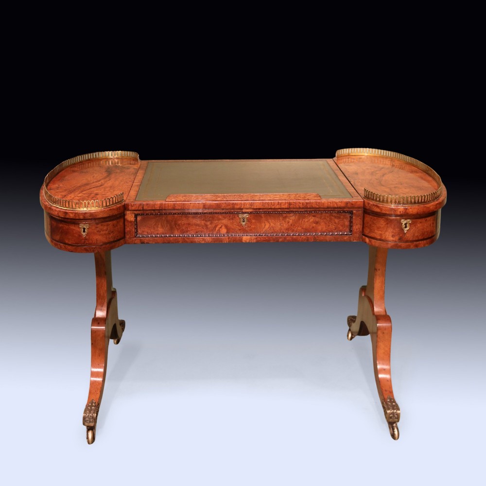 An early 19th century Regency burr oak writing table featuring a green leather inset top, brass gallery rails on raised side platforms, and trestle legs with brass castors.