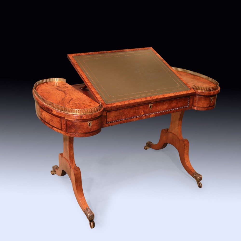 An early 19th century Regency burr oak writing table featuring a green leather inset top, brass gallery rails on raised side platforms, and trestle legs with brass castors. View II