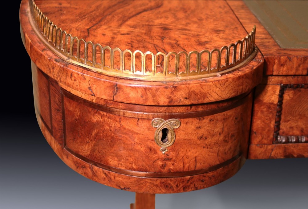 An early 19th century Regency burr oak writing table featuring a green leather inset top, brass gallery rails on raised side platforms, and trestle legs with brass castors. View IV