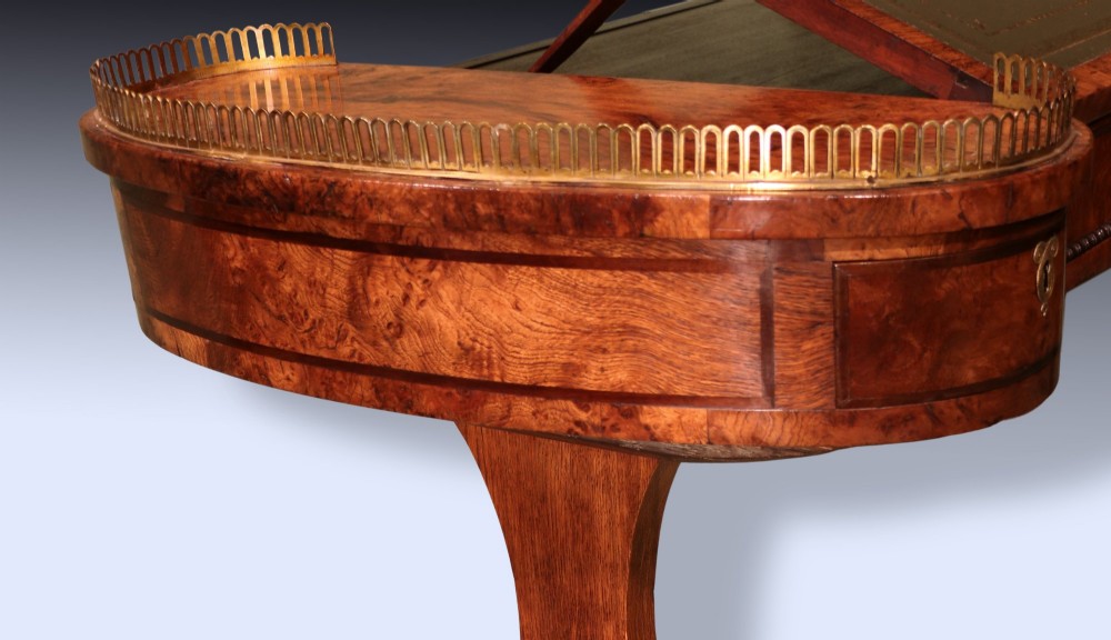 An early 19th century Regency burr oak writing table featuring a green leather inset top, brass gallery rails on raised side platforms, and trestle legs with brass castors. View V