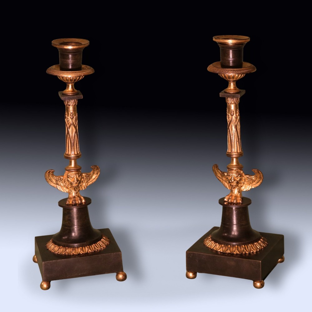 A pair of 19th-century French Empire style candlesticks featuring patinated bronze columns supported by gilded ormolu winged lions on square bases.