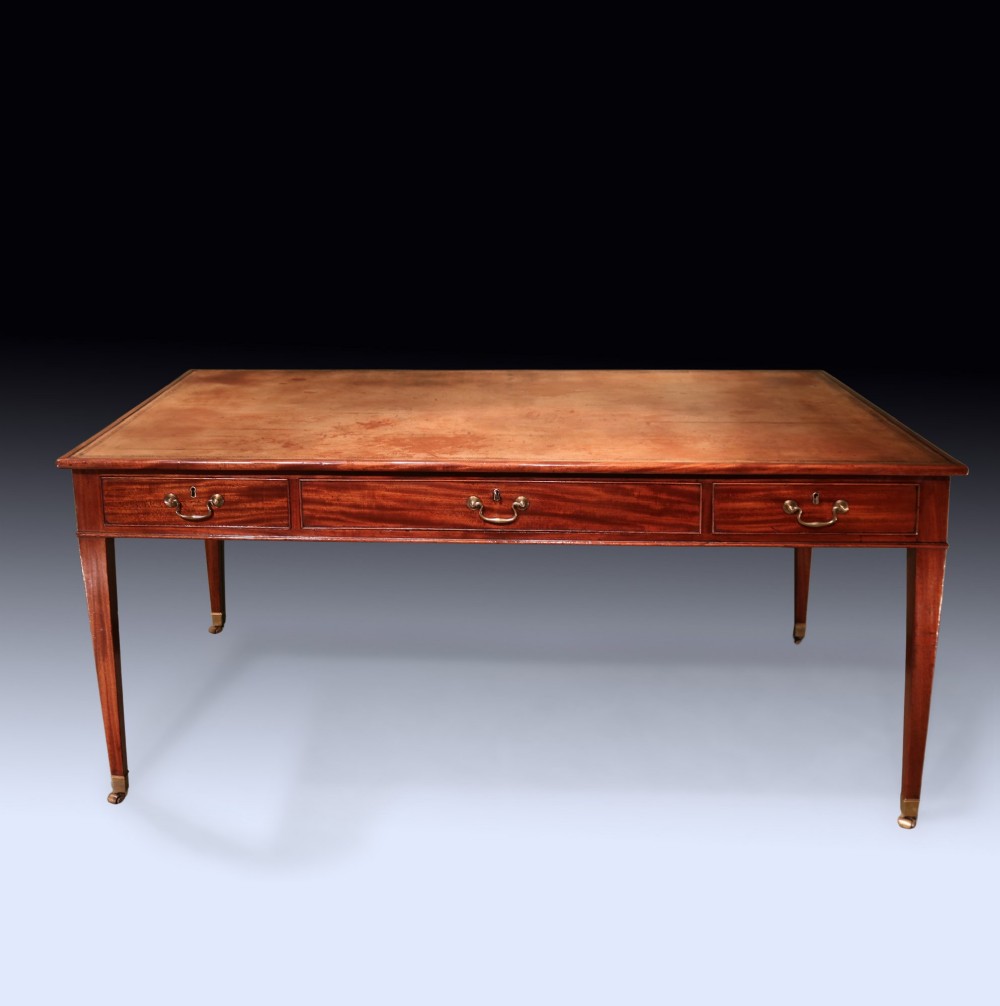 An antique George III mahogany writing table featuring an inset leather top, three frieze drawers with brass handles, and slender square tapered legs on castors.