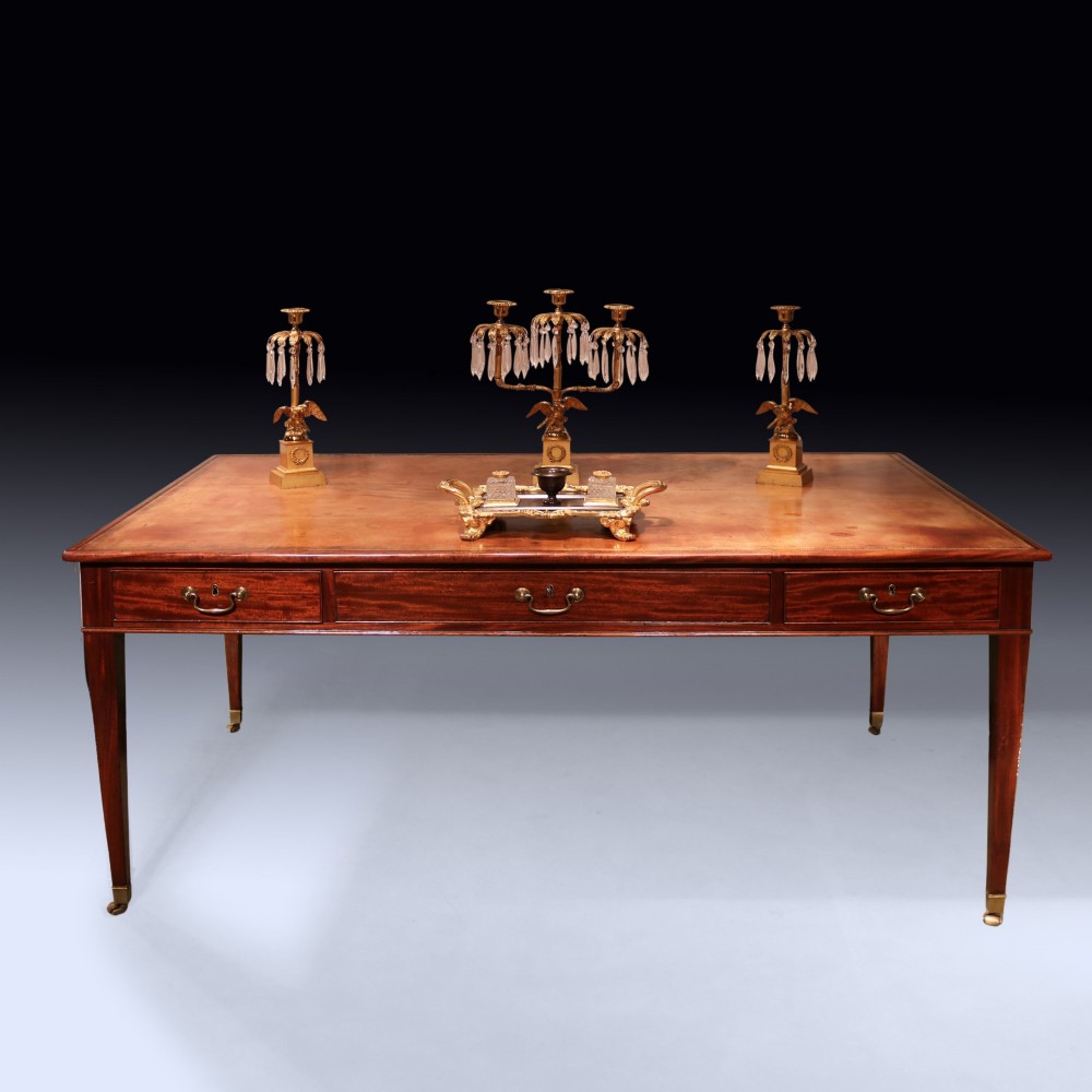 An antique George III mahogany writing table featuring an inset leather top, three frieze drawers with brass handles, and slender square tapered legs on castors. View IV