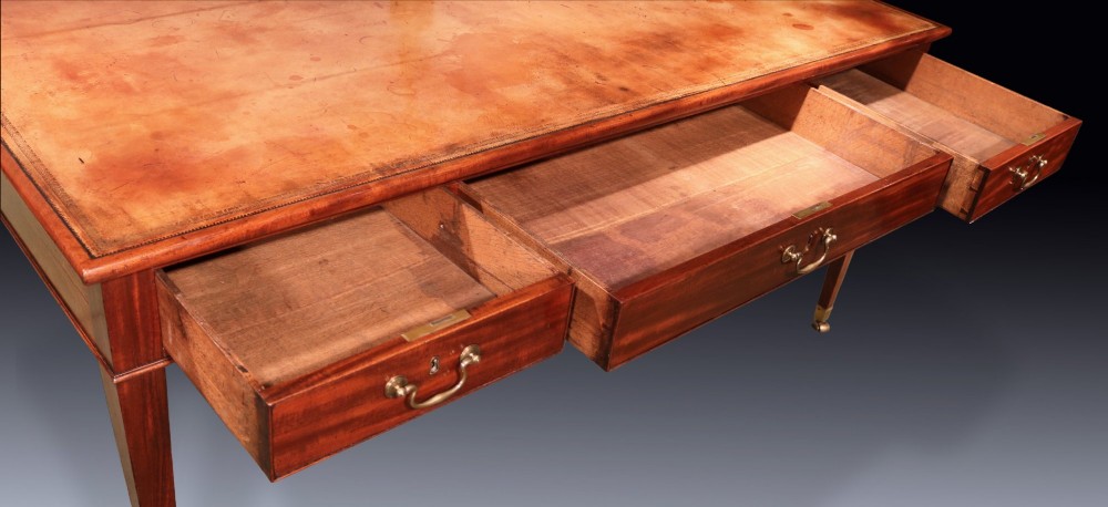 An antique George III mahogany writing table featuring an inset leather top, three frieze drawers with brass handles, and slender square tapered legs on castors. View V