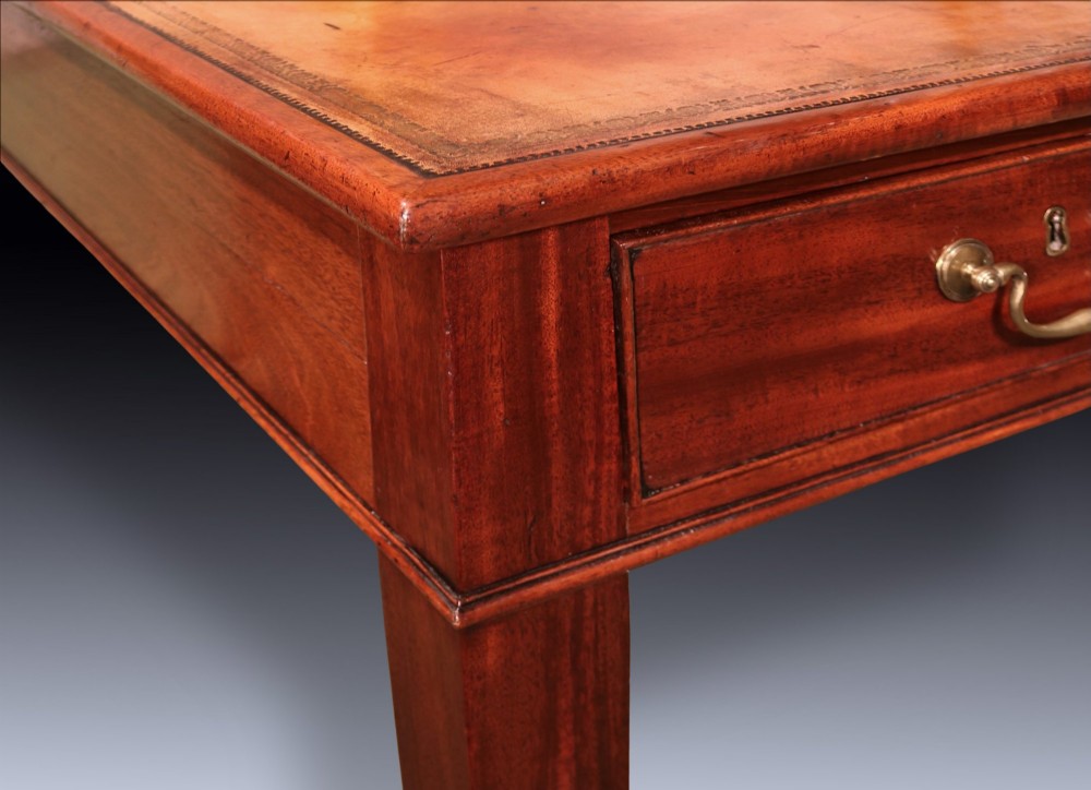 An antique George III mahogany writing table featuring an inset leather top, three frieze drawers with brass handles, and slender square tapered legs on castors. View VI