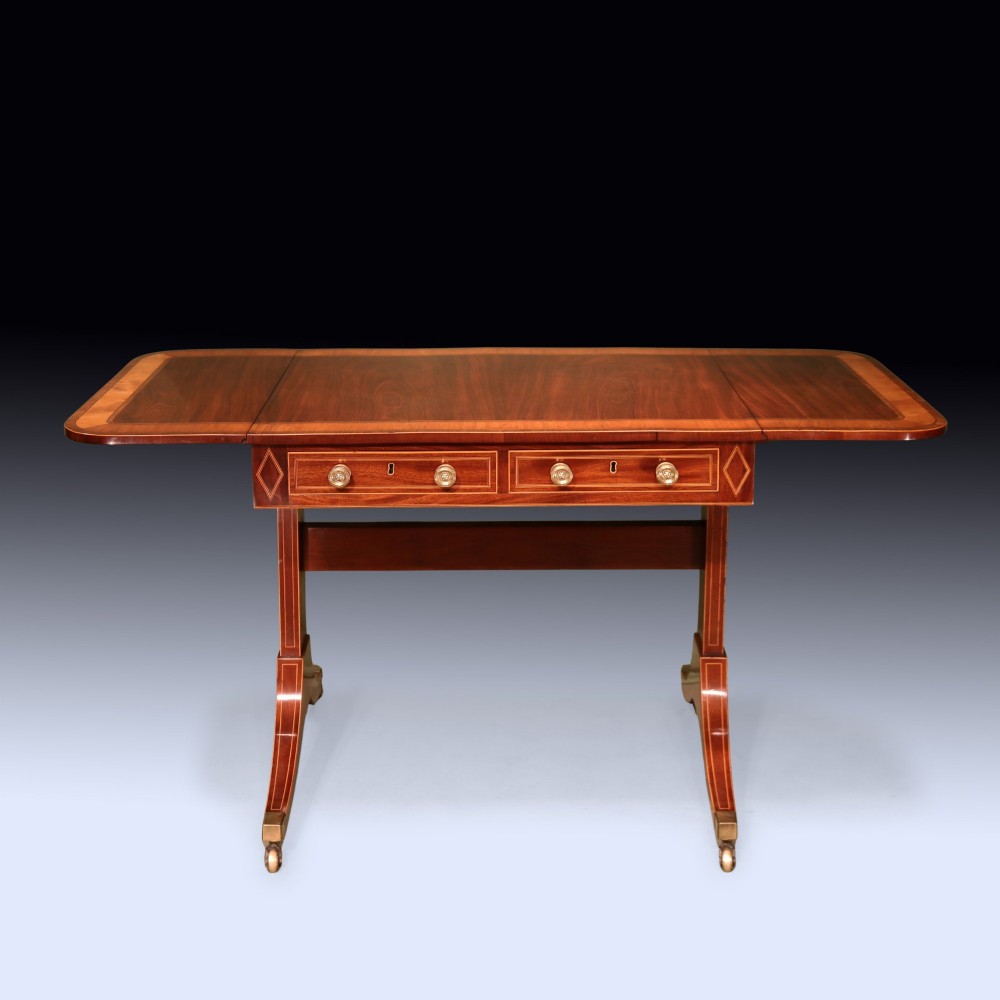 A George III mahogany sofa table featuring satinwood crossbanding, drop-leaf ends, two frieze drawers, and splayed legs on brass castors.
