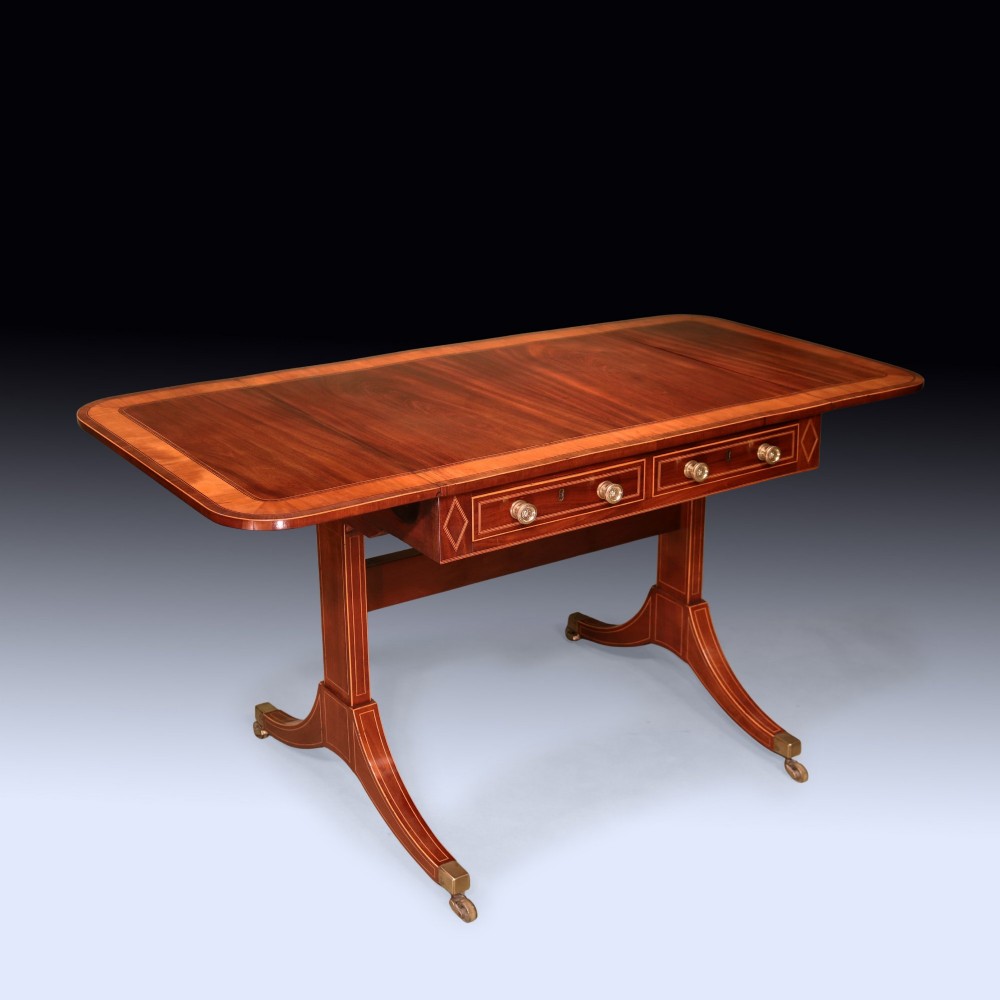 A George III mahogany sofa table featuring satinwood crossbanding, drop-leaf ends, two frieze drawers, and splayed legs on brass castors. View II