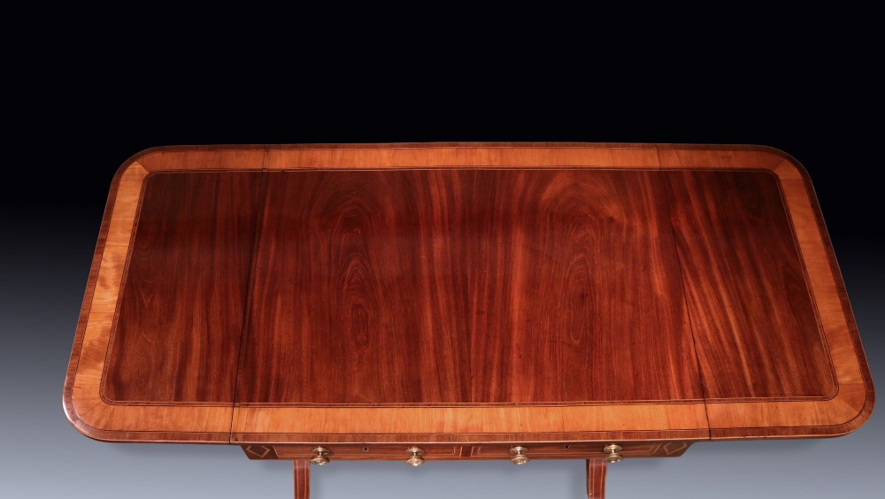 A George III mahogany sofa table featuring satinwood crossbanding, drop-leaf ends, two frieze drawers, and splayed legs on brass castors. View III