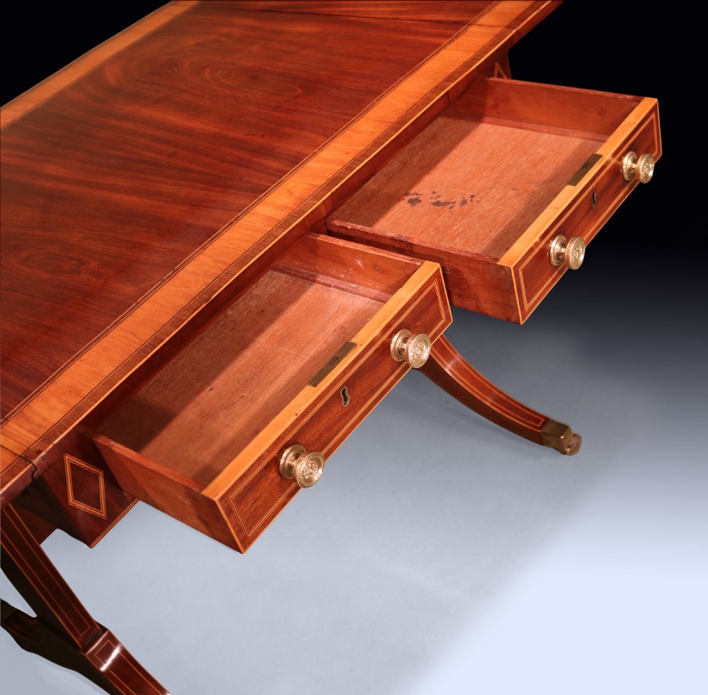 A George III mahogany sofa table featuring satinwood crossbanding, drop-leaf ends, two frieze drawers, and splayed legs on brass castors. View IV