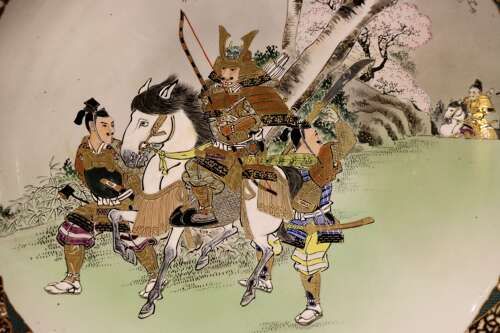fig 1. A large circular Japanese Kutani porcelain charger featuring hand-painted samurai warriors on horseback under a cherry blossom tree, surrounded by a decorative green and gold patterned rim.