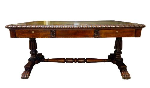 fig 1. A George IV mahogany partners writing table featuring a patterned top edge, three frieze drawers, carved pedestal supports, a turned stretcher, and claw feet.