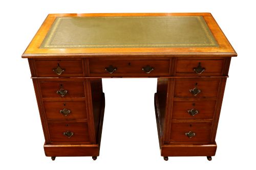 fig 2. A small Edwardian walnut pedestal desk featuring a green leather top with gold tooling, nine brass-handled drawers, fluted side details, and castors at the base.