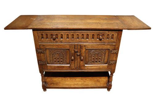 fig 2. A warm brown carved oak side cabinet featuring drop-leaf top extensions, arcaded carved cupboard doors, turned legs, and a lower pot board shelf.