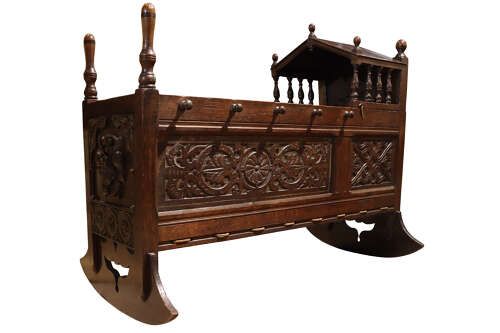 fig 1. A dark carved oak rocking cradle featuring a hooded top with turned spindles, carved relief side panels, and curved rockers at the base.
