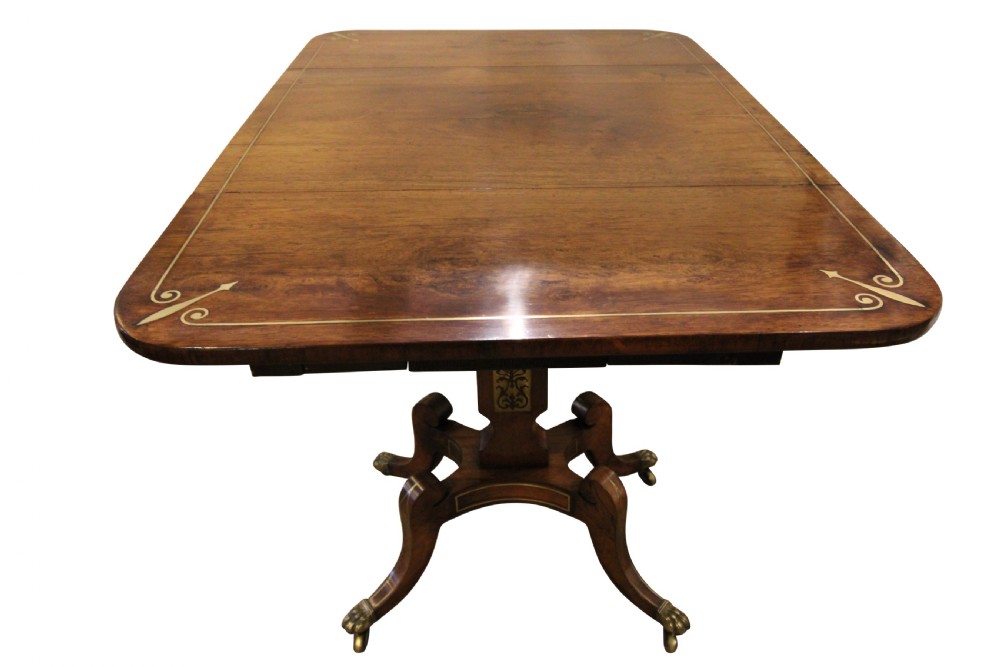 A Regency period rosewood drop-leaf work table featuring two drawers with circular brass knobs, supported by a central pedestal and four curved legs with brass lion paw castors. View III