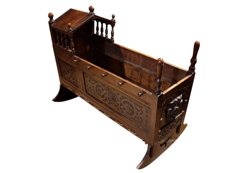 A dark oak hooded rocking cradle featuring carved geometric panels, turned side spindles, tall corner finials, and curved rocking bases.
