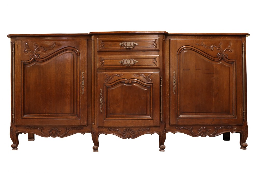A French Louis XV style warm brown oak sideboard featuring three carved panel doors, two central drawers, brass hardware, and cabriole feet.