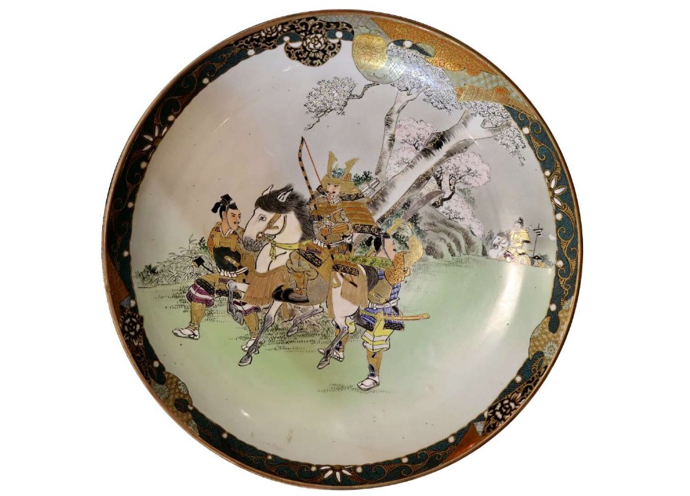 A large circular Japanese Kutani porcelain charger featuring hand-painted samurai warriors on horseback under a cherry blossom tree, surrounded by a decorative green and gold patterned rim.
