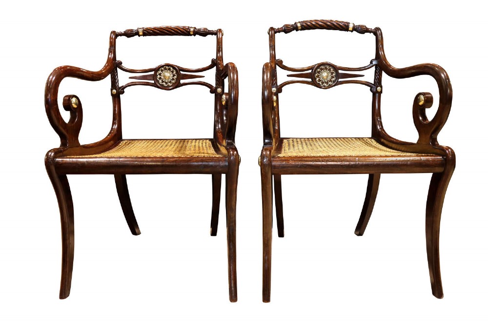 A pair of dark mahogany Regency armchairs featuring swept sabre legs, woven cane seats, rope-twist top rails, and central brass rosette mounts on the backrest splats.