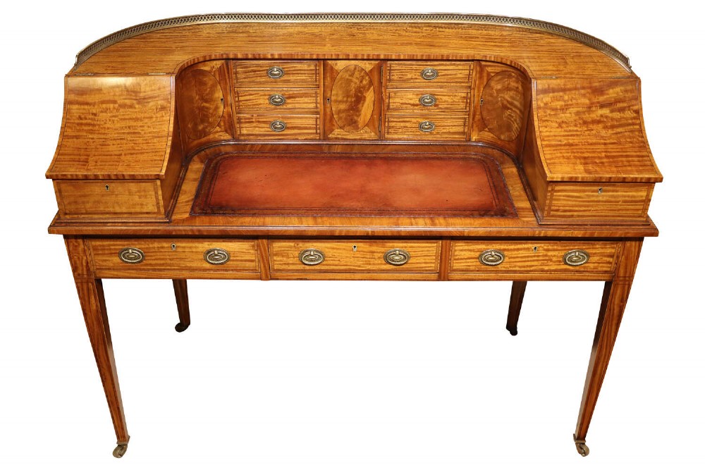An Edwardian satinwood Carlton House desk featuring a curved top gallery with small drawers, marquetry inlay, a leather writing surface, and tapered legs on brass castors.