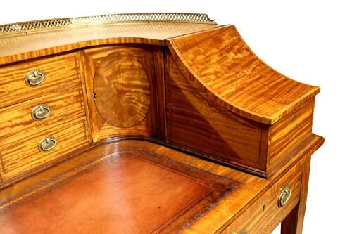 An Edwardian satinwood Carlton House desk featuring a curved top gallery with small drawers, marquetry inlay, a leather writing surface, and tapered legs on brass castors. View IV
