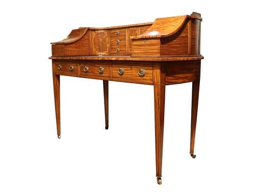 An Edwardian satinwood Carlton House desk featuring a curved top gallery with small drawers, marquetry inlay, a leather writing surface, and tapered legs on brass castors. View V