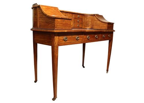 An Edwardian satinwood Carlton House desk featuring a curved top gallery with small drawers, marquetry inlay, a leather writing surface, and tapered legs on brass castors. View VI