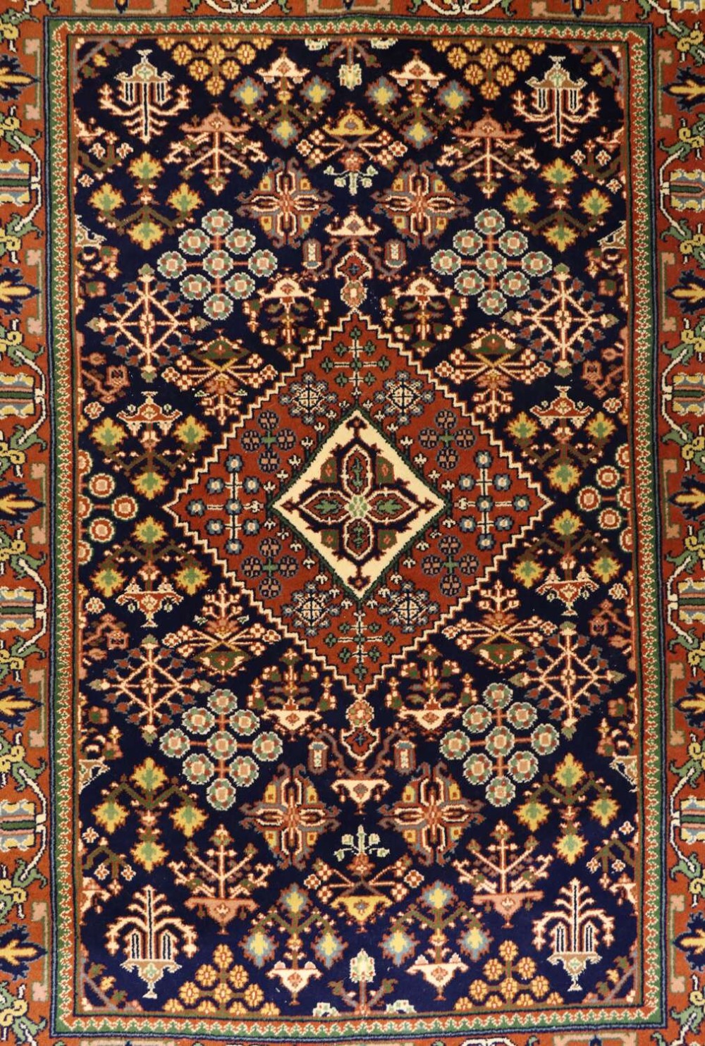 A hand-knotted Persian wool area rug featuring a central diamond medallion surrounded by geometric tribal motifs in shades of navy blue, terracotta red, green, and cream.