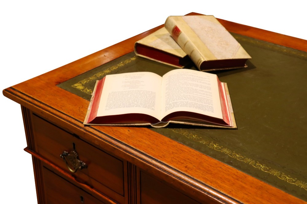 A small Edwardian walnut pedestal desk featuring a green leather top with gold tooling, nine brass-handled drawers, fluted side details, and castors at the base. View V