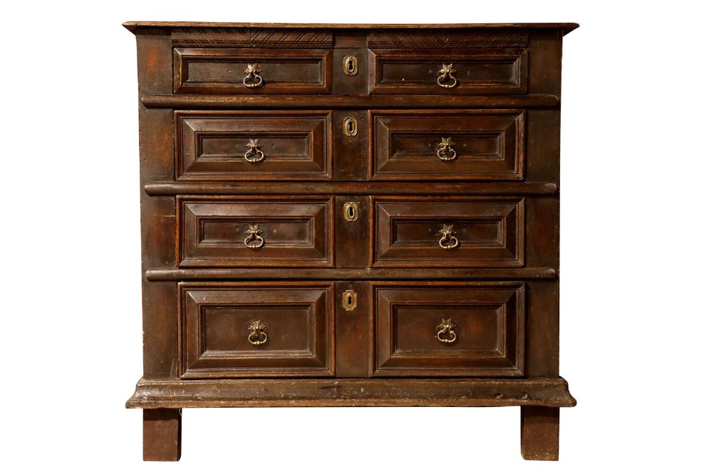 A dark oak Charles II chest of drawers featuring geometric panelled fronts, brass teardrop handles, and keyhole escutcheons.