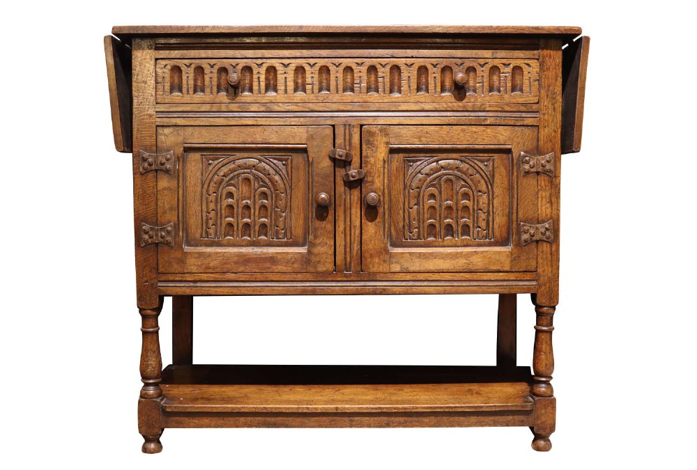 A warm brown carved oak side cabinet featuring drop-leaf top extensions, arcaded carved cupboard doors, turned legs, and a lower pot board shelf.