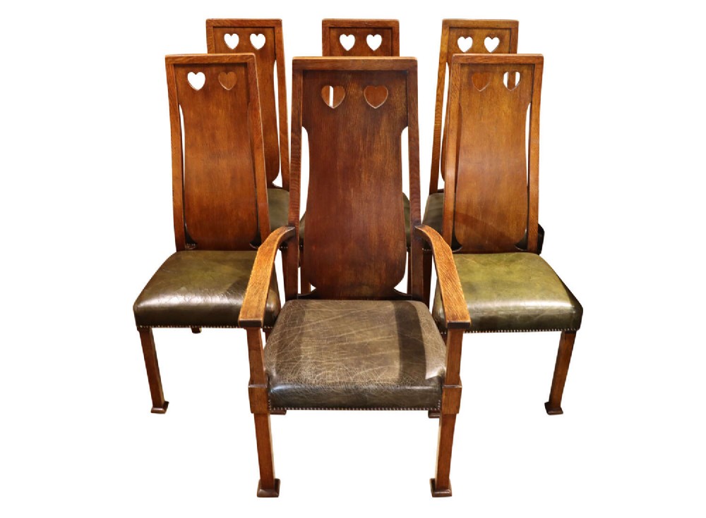 A set of six Arts and Crafts solid oak dining chairs featuring tall backsplats with heart-shaped cutouts and green leather seats with brass studding.